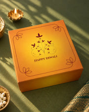 Closed saffron-orange box printed Happy Diwali beside a lit brass diya — Aaradhya Diwali gift hamper
