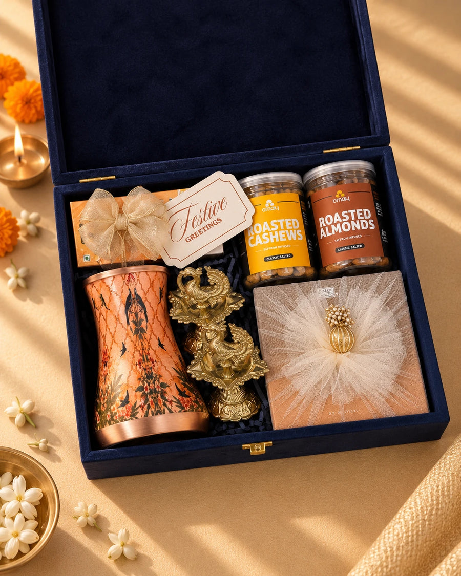 Open navy velvet box with a Meena-print copper glass, brass peacock diyas and saffron nut jars — Aarambh Diwali gift hamper