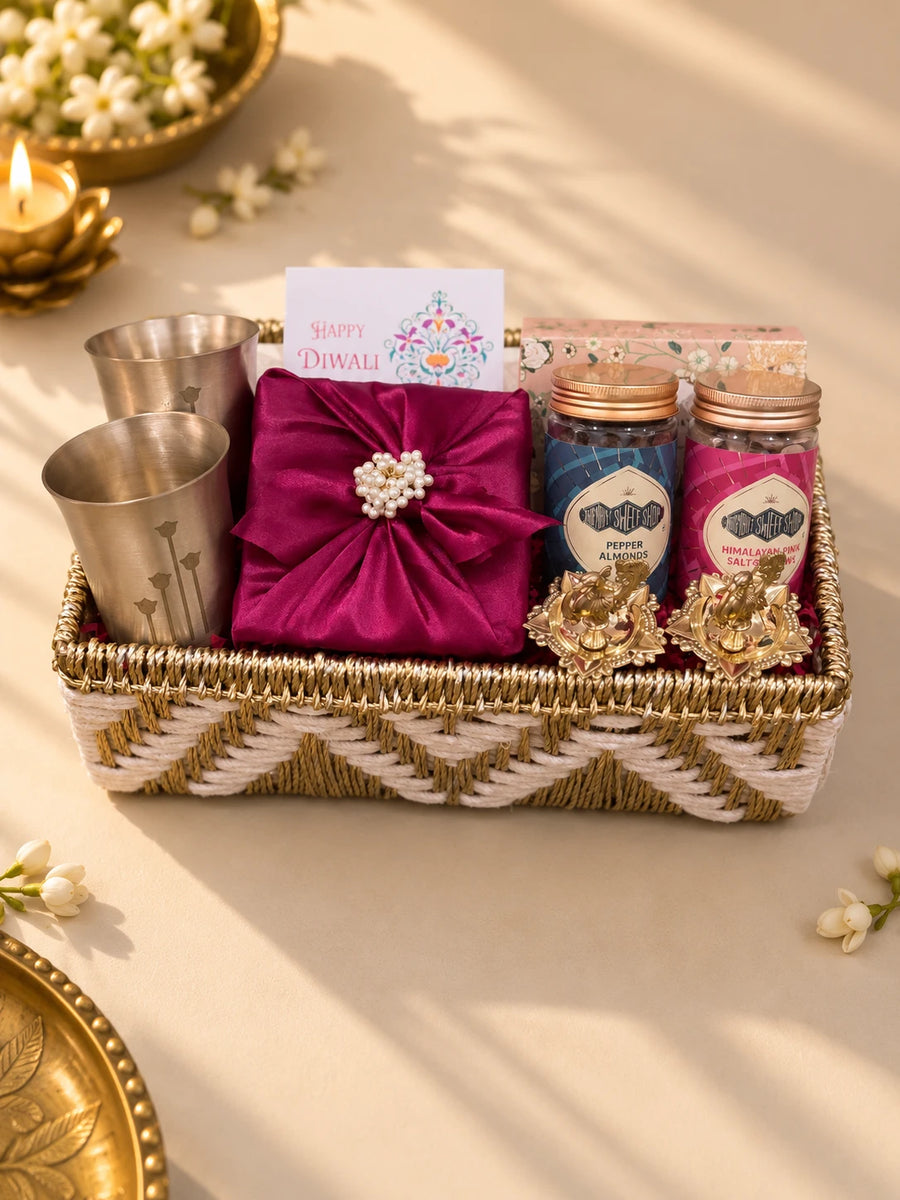 Woven ivory tray with two matte brass glasses, a magenta silk-wrapped box, nut jars and diyas — Tattva Diwali gift hamper